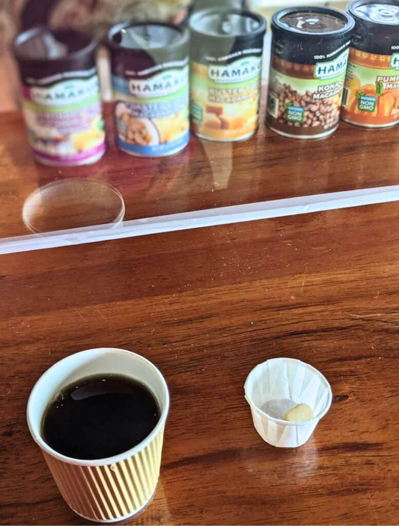 Small cup of black coffee served beside a sample of macadamia nut on a wooden counter with cans labeled "HAMAKUA" in flavors including "Kona Coffee Macadamias" and "Pumpkin Macadamias" displayed behind. Tasting local macadamia products is a delicious stop near Hawi for visitors exploring things to do Hawi.