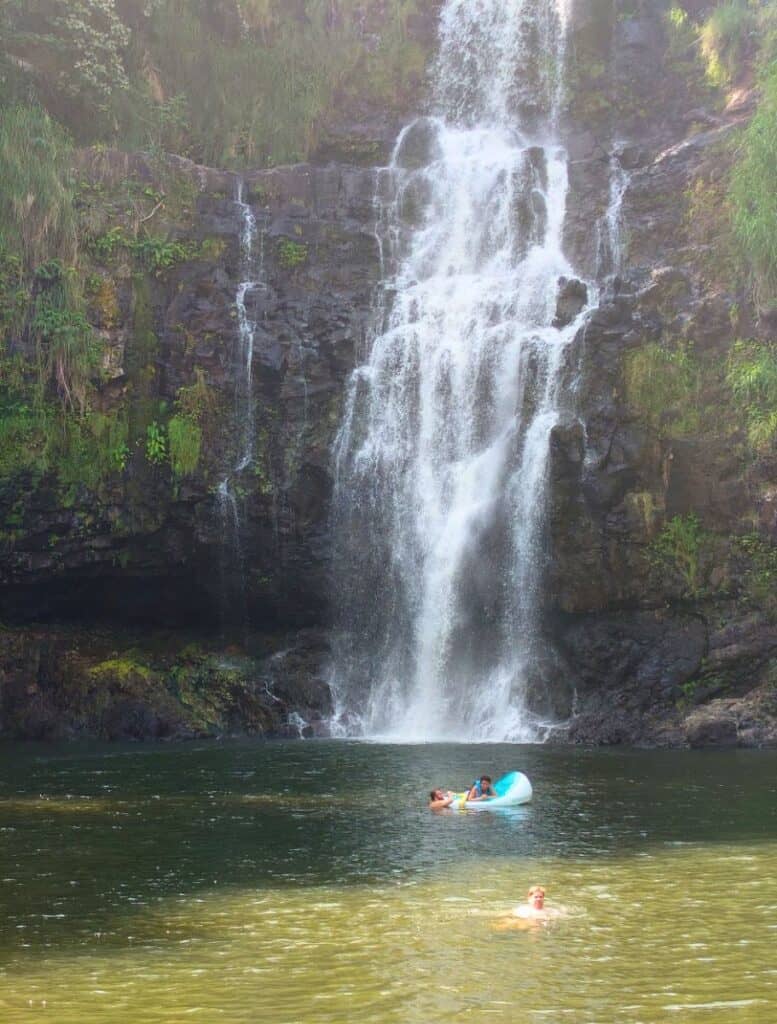 Tall waterfall cascading down a dark rocky cliff into a calm pool where people swim and float on an inflatable, showcasing one of the big island waterfalls you can swim in surrounded by lush green vegetation. Sunlight filters through the mist as a swimmer in the foreground wades through the shallow water near the base of the falls.