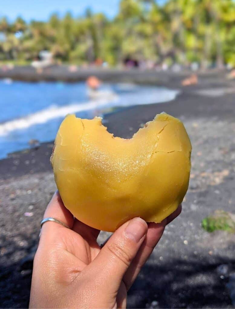 my hand holding a lilikoi malasada with one bite taken out of it. punalu'u black sand beach in the background