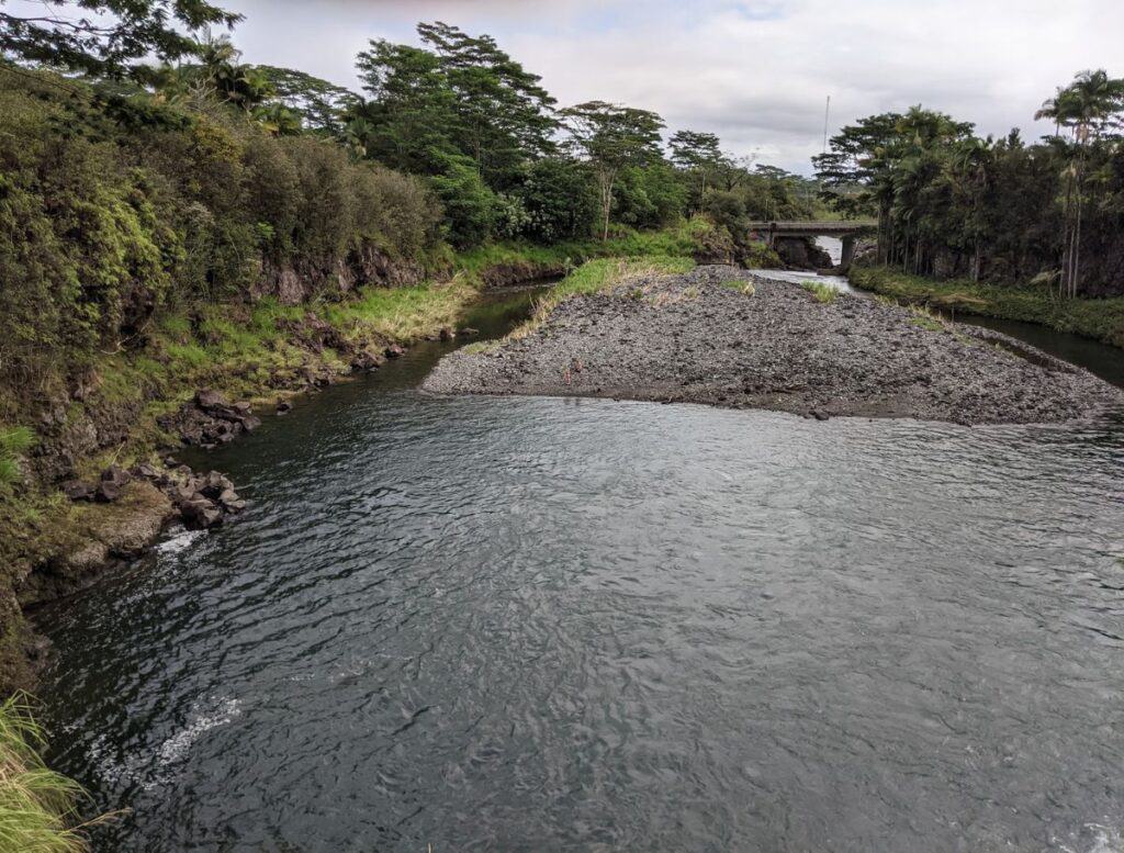 Wide river pool bordered by rocky banks and lush tropical trees with a small bridge in the background, showing a calm swimming area connected to big island waterfalls you can swim in. The gently flowing water and open space create a relaxed spot for swimming near the falls upstream.