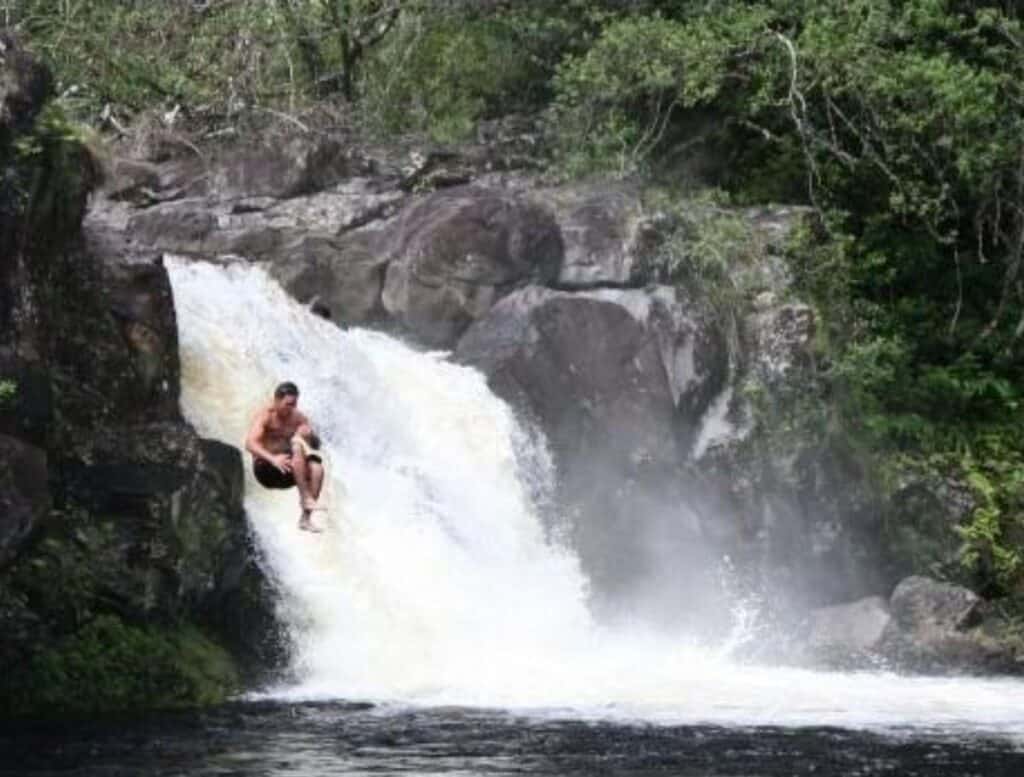 Person sliding down a smooth natural rock chute beside a small waterfall into a deep pool, highlighting an adventurous spot among big island waterfalls you can swim in surrounded by rugged rocks and dense greenery. White water rushes over the dark stone as the swimmer drops into the calm basin below.