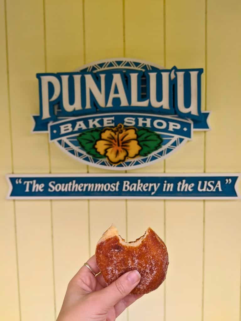 Hand holding a partially eaten pastry in front of a yellow wall with a sign that reads "Punaluu Bake Shop" and "The Southernmost Bakery in the USA." Stopping here for a sweet local treat is a fun food stop to include in a big island itinerary 4 days trip.