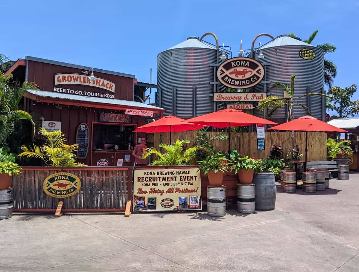 Kona brewing co front with two large silos, 3 red umbrellas, and a growlers shack