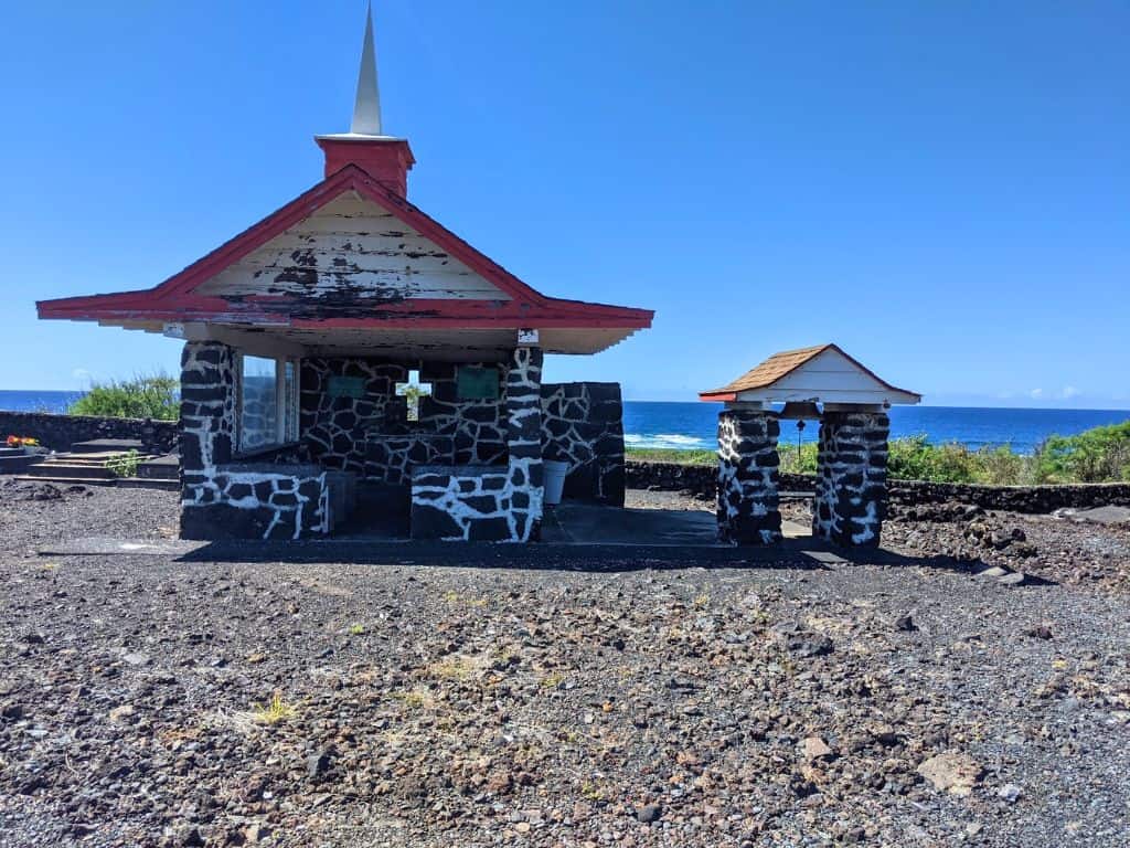 Hokuloa Church at Punalu'u Black Sand Beach, an open air stone chapel with a bell next to it and water view in background