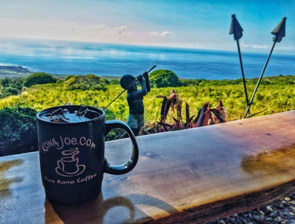Black mug that reads "KonaJoe.com Pure Kona Coffee" sits on a wooden counter overlooking green coffee fields and the blue ocean in the distance. The scenic farm view shows a classic Kona coffee experience often featured in a Big Island travel guide.
