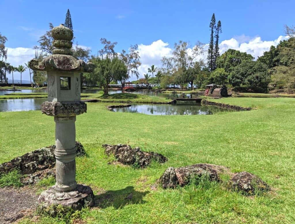 Japanese style stone lantern in a manicured garden with ponds palm trees and a red bridge in the distance under a bright blue sky in Hilo Hawaii. Peaceful gardens like this offer scenic walking paths and are one of the relaxing things to do in Hilo Hawaii.