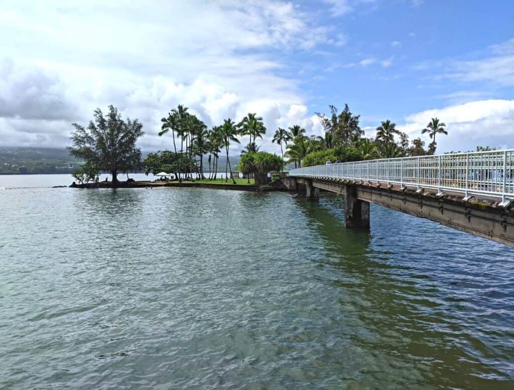 White pedestrian bridge stretching over calm water toward a small palm tree covered island in Hilo Hawaii with mountains and clouds in the distance. Walking across scenic coastal parks like this is a peaceful activity and one of the relaxing things to do in Hilo Hawaii.