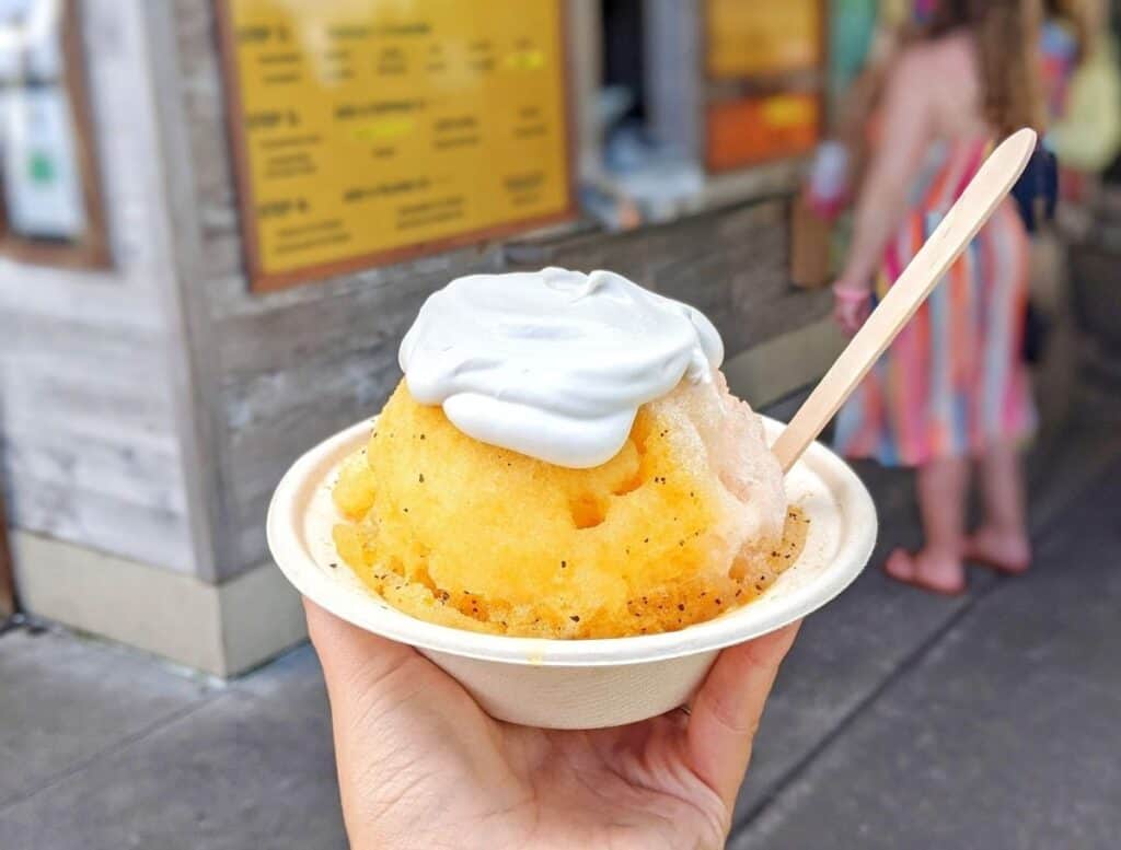 Hand holding a bowl of bright yellow shave ice topped with a scoop of white ice cream and a wooden spoon in front of a small roadside stand. This classic Hawaiian dessert is a refreshing treat to cool down while exploring Hilo.