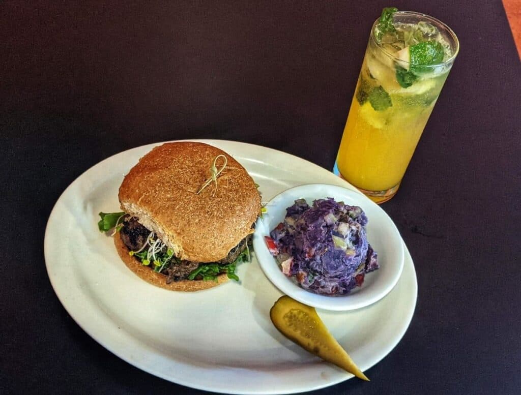 Plate with a gourmet burger on a whole grain bun topped with greens served alongside a scoop of purple potato salad and a pickle spear with a tall citrus mint drink. This colorful meal highlights a fresh local dining experience in Hilo Hawaii.