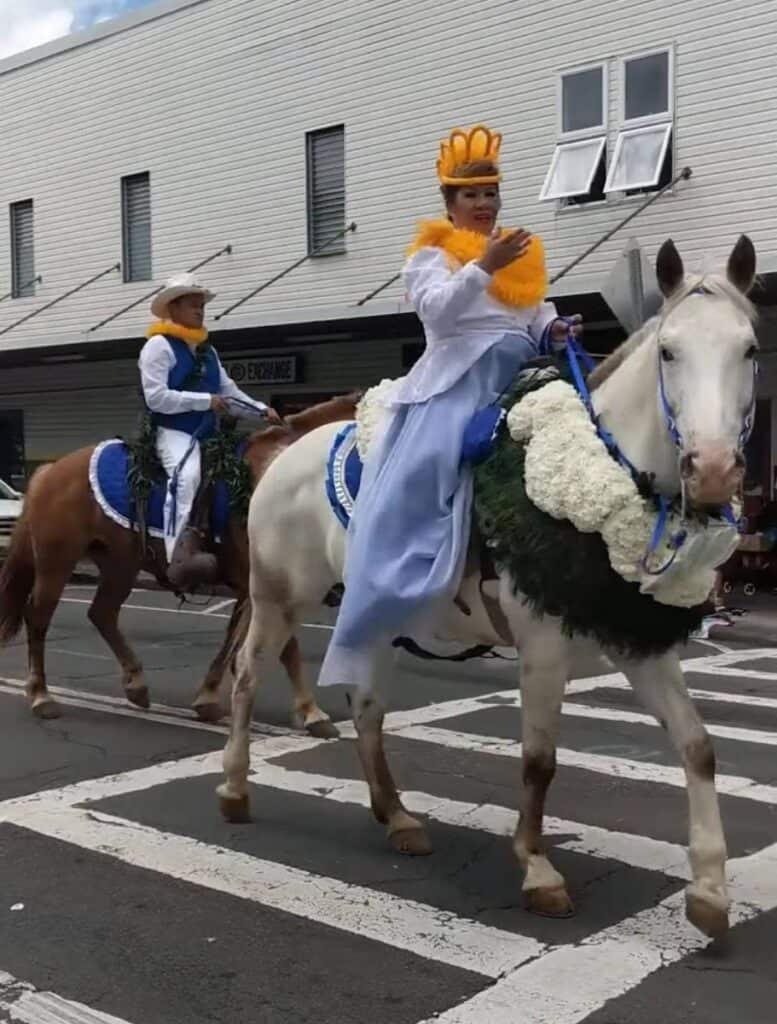 Woman wearing a crown and yellow lei riding a decorated horse during a local parade in Hilo Hawaii with another rider following behind. Colorful cultural parades and festivals like this are lively events and one of the unique things to do in Hilo Hawaii.