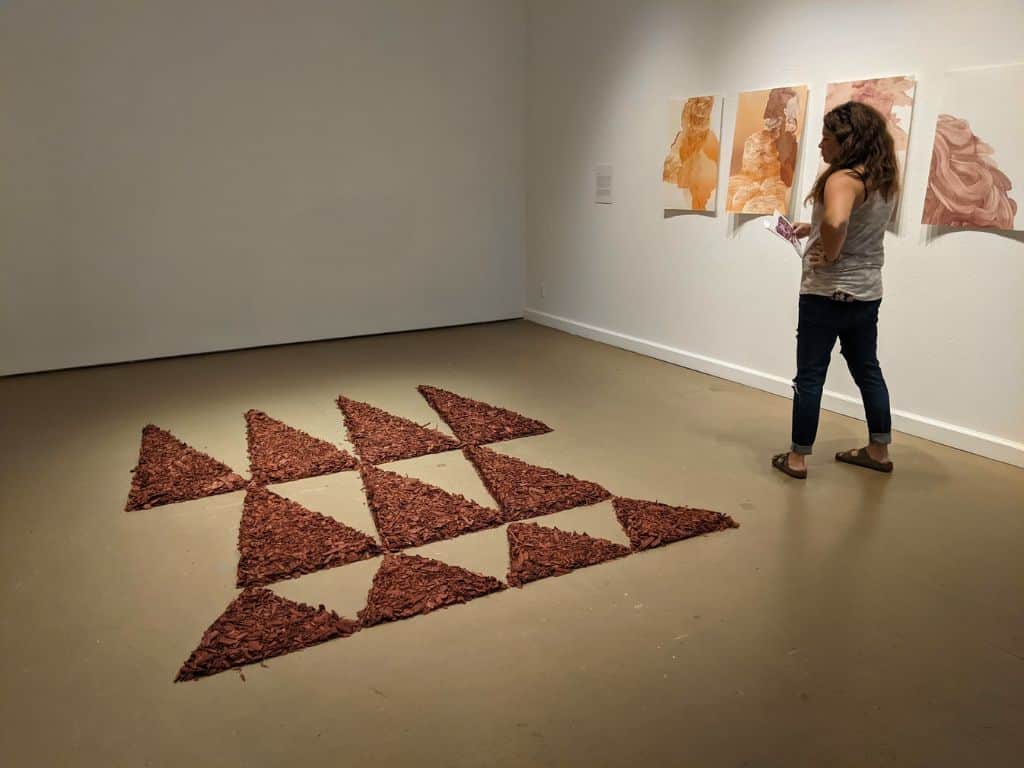 Woman viewing artwork in a modern gallery space with abstract triangular installation pieces arranged on the floor and warm toned paintings on the wall in Hilo Hawaii. Visiting local art galleries to experience contemporary Hawaiian art is one of the creative things to do in Hilo Hawaii.