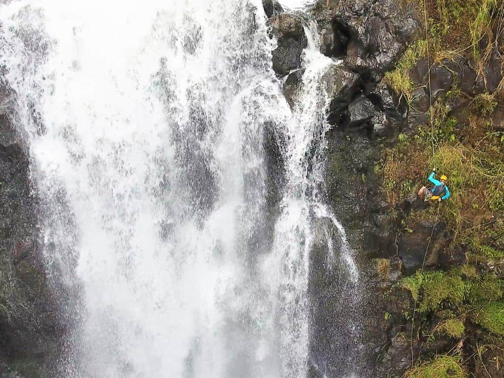 Person wearing a helmet rappelling down a steep cliff beside a powerful waterfall with rushing white water and dark volcanic rock in Hilo Hawaii. Waterfall rappelling through lush tropical scenery is an adventurous outdoor activity and one of the thrilling things to do in Hilo Hawaii.