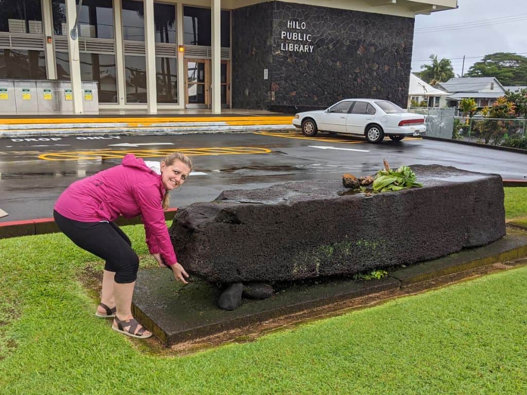 Woman in a pink jacket posing as if lifting a large stone slab display outside the "Hilo Public Library" building in Hilo Hawaii with a sign visible on the wall. Visiting local landmarks and historic sites like the library grounds is one of the interesting things to do in Hilo Hawaii.