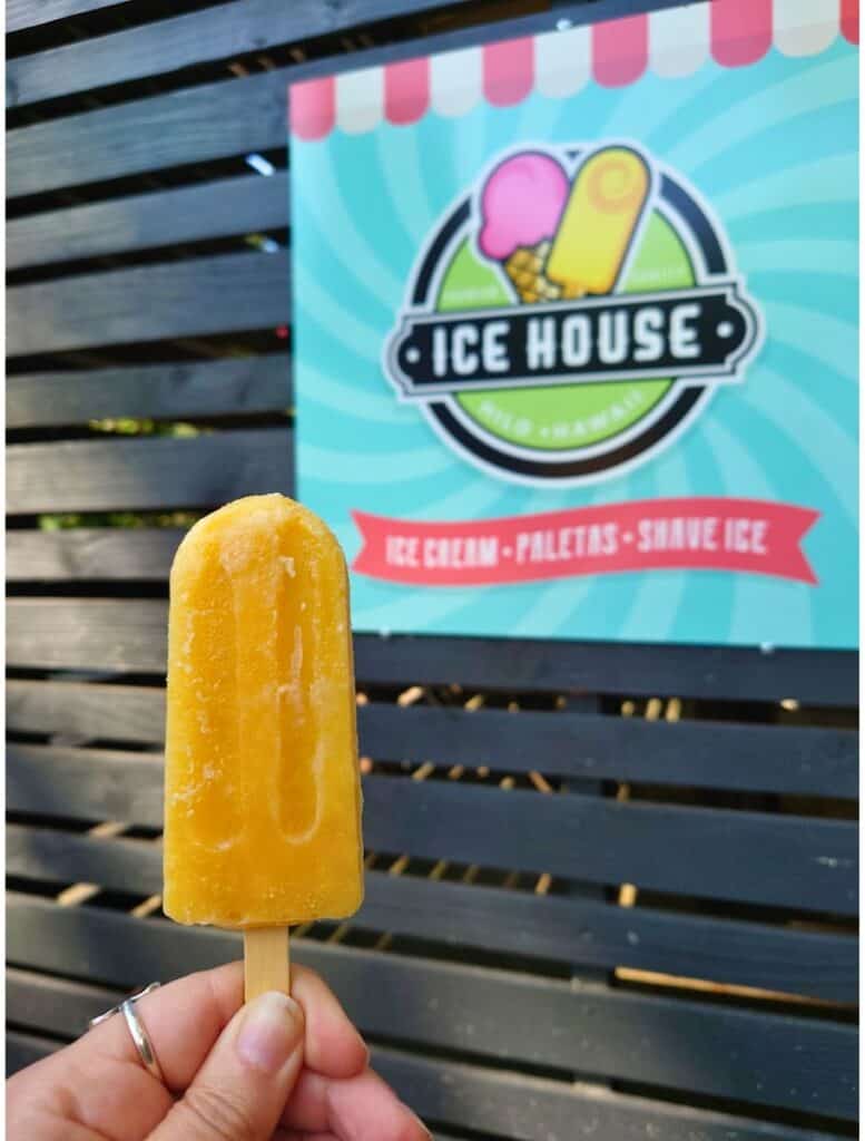 Hand holding a bright orange fruit popsicle on a wooden stick in front of a sign reading "Ice House Ice Cream Paletas Shave Ice". This refreshing frozen treat is a popular stop for cooling off while exploring Hilo.