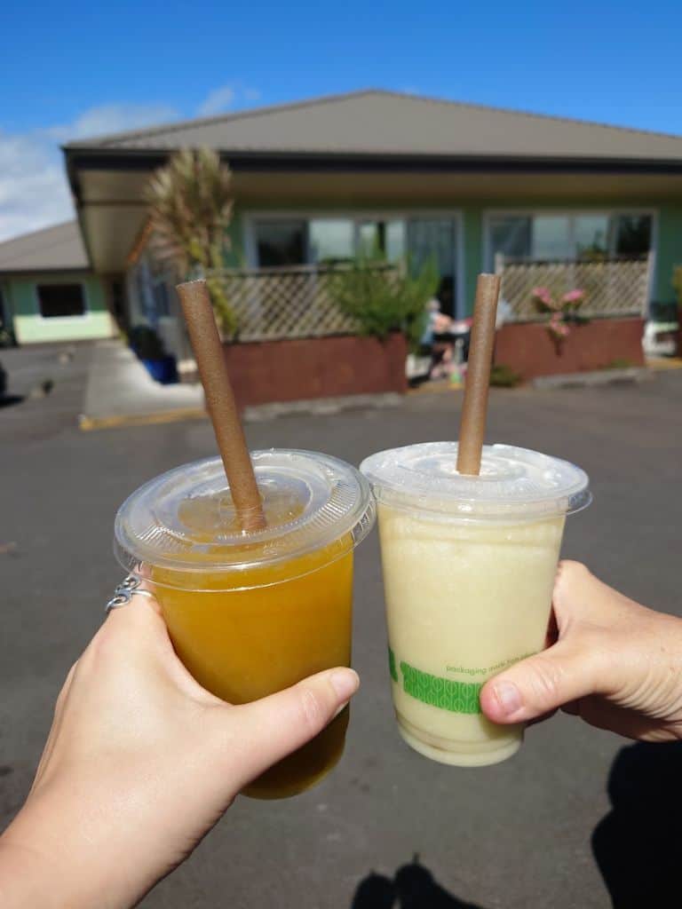 Two hands holding iced tropical drinks in clear cups with straws in front of a small local shop on a sunny day. Refreshing sugarcane juice is a refreshing treat to cool off in Hilo Hawaii.