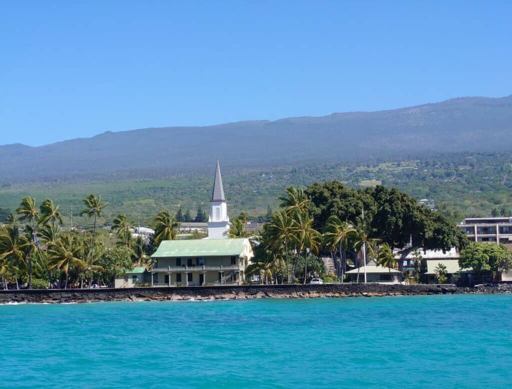 White seaside church with a tall steeple surrounded by palm trees along the bright turquoise coastline of the Big Island. Green hills rise in the background behind the historic shoreline scene often featured in a Big Island travel guide.