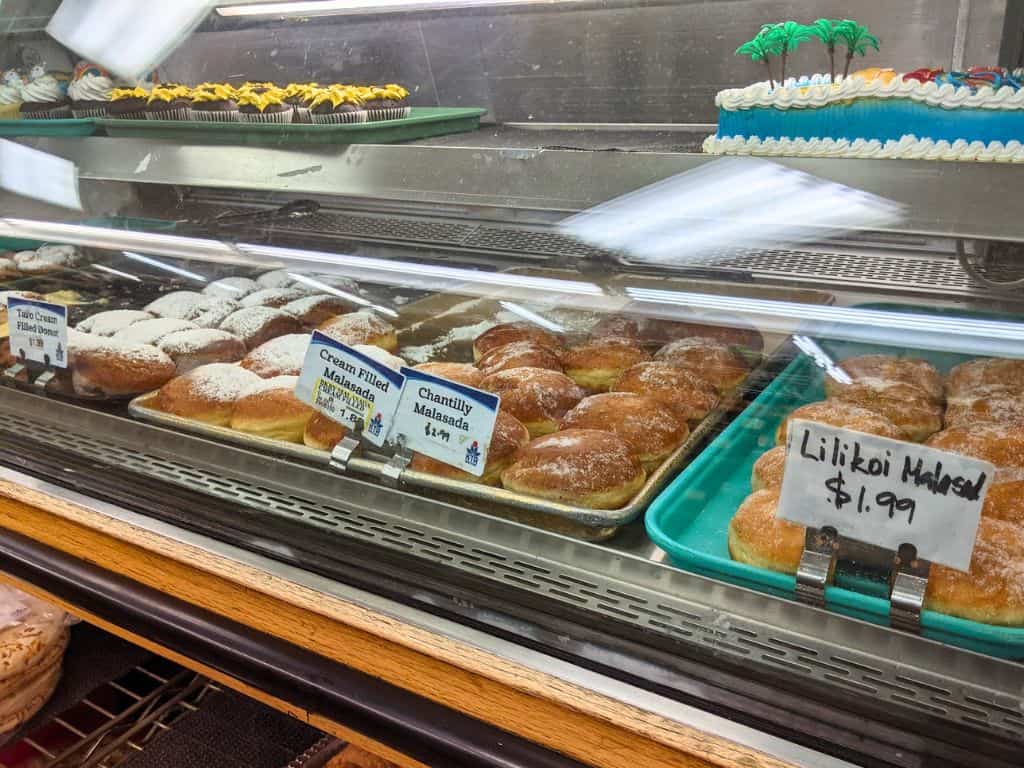 Malasadas in glass display at KTA Super Store. Flavors labeled: taro cream filled donut, cream filled malasada, chantilly malasada, and lilikoi malasada