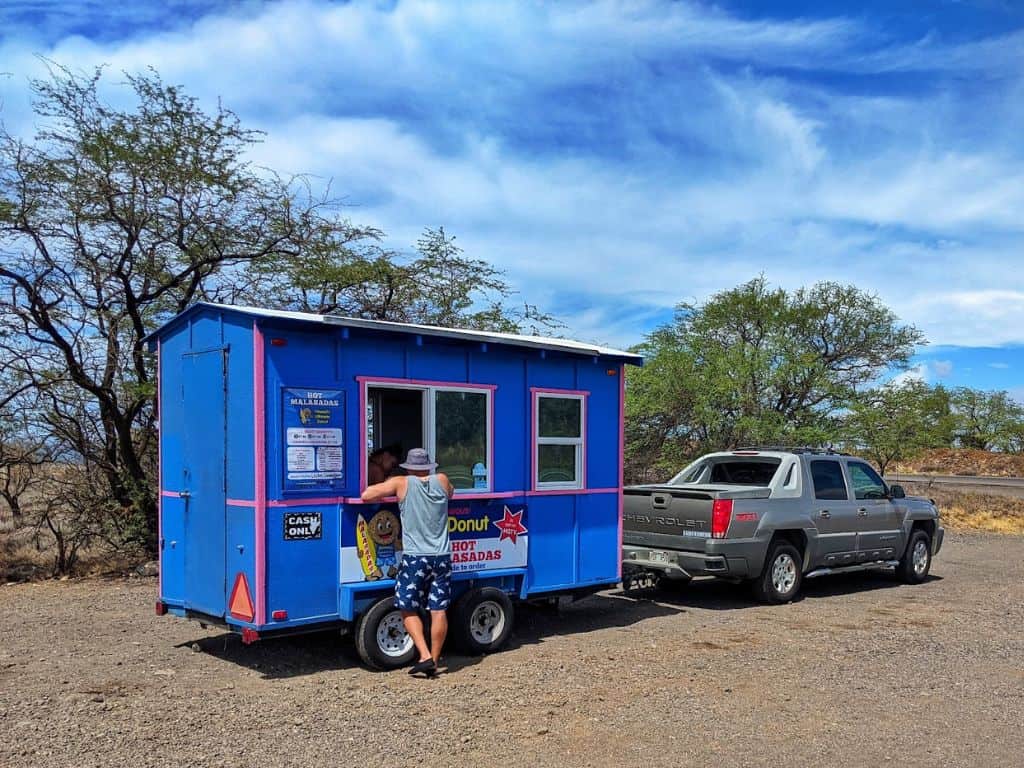 person ordering from Manuela's Malasadas blue food truck trailer in gravel parking lot