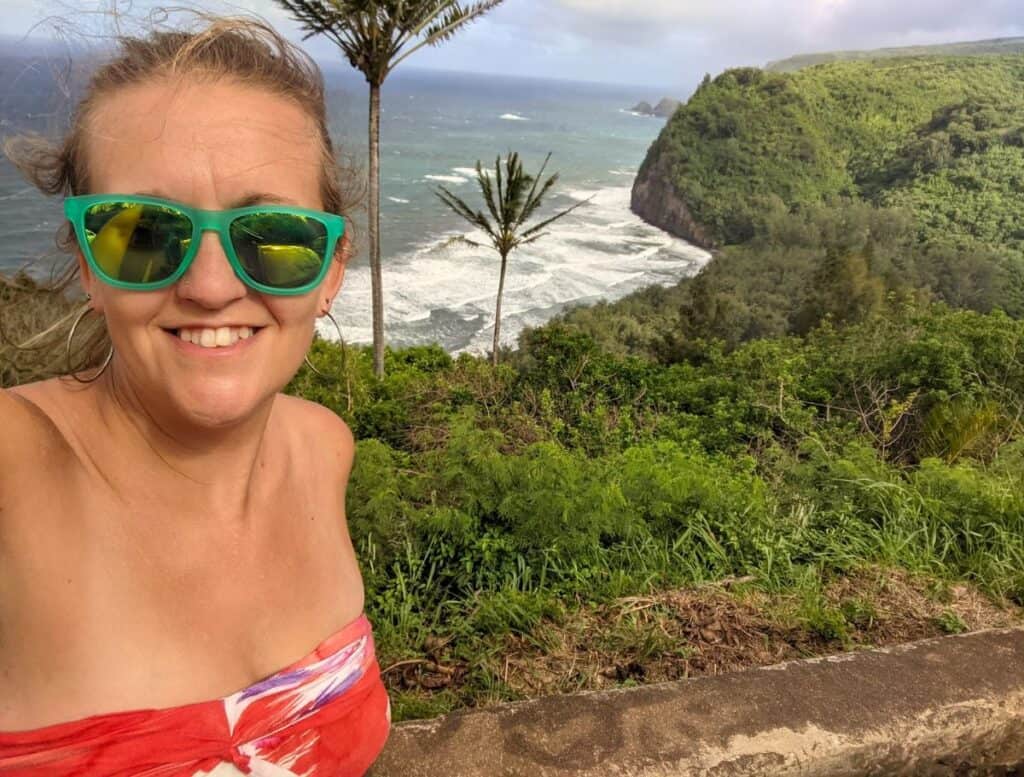 Woman wearing green sunglasses smiles in a selfie at a scenic overlook above a rugged coastline with steep green cliffs and waves crashing below. Tropical viewpoint near Hawi where visitors stop for photos while exploring things to do Hawi along the northern Big Island coast.