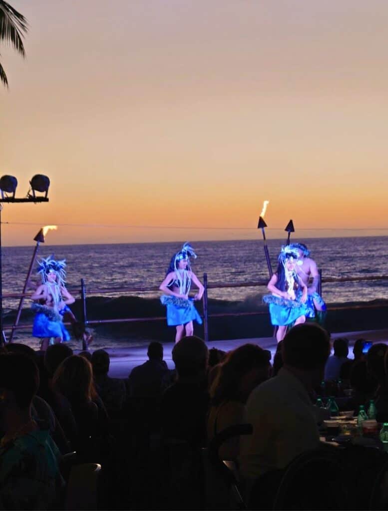 Luau performers in blue skirts and feather headdresses dancing on stage with ocean in background