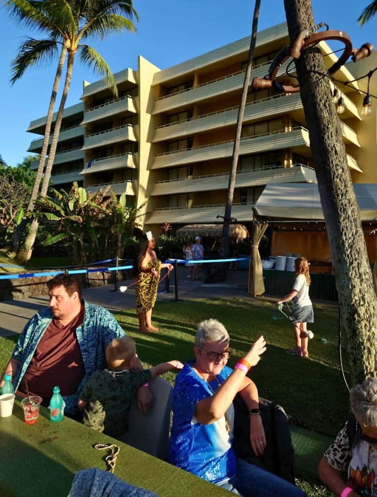 woman shades her eyes with hand from sun as she is sitting at table surrounded by family members. royal kona resort building in background