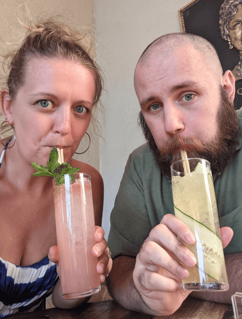 Two travelers sit at a table in Waikoloa sipping colorful tropical cocktails through straws, one pink drink garnished with fresh mint and one pale drink with a cucumber slice. The casual vacation moment highlights enjoying local drinks and relaxing together, one of the many things to do Waikoloa visitors experience while dining out.