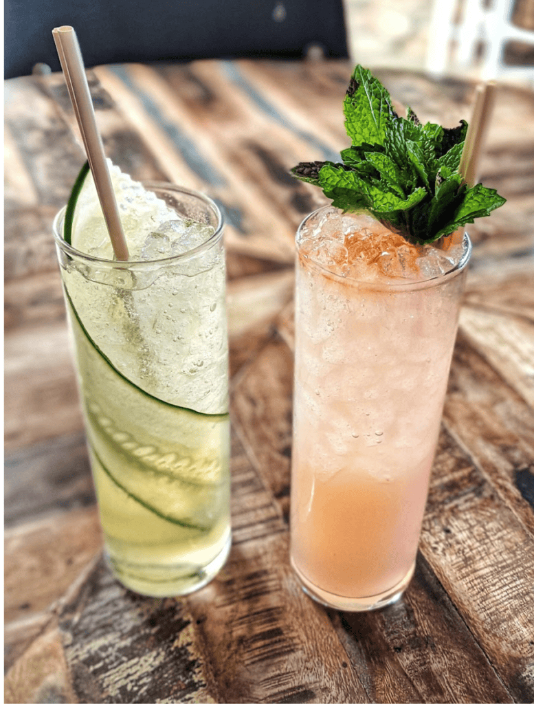 Two tall refreshing cocktails on a wooden table. One drink is garnished with thin cucumber slices and the other with fresh mint leaves. Enjoying tropical cocktails at a local bar or restaurant is one of the relaxing things to do Waikoloa after a day at the beach.