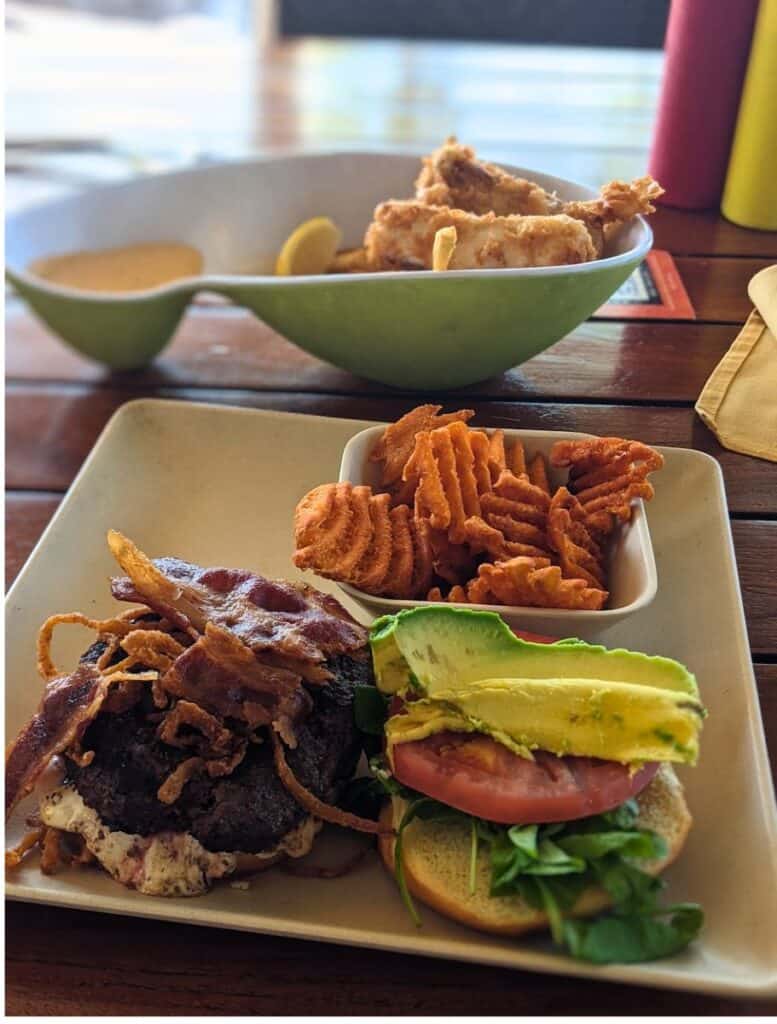 Burger topped with avocado, tomato, bacon, and crispy onions served with waffle fries and fried fish on a wooden table at a casual restaurant. Trying local restaurants and comfort food near the beach is one of the tasty things to do Waikoloa between outdoor adventures.