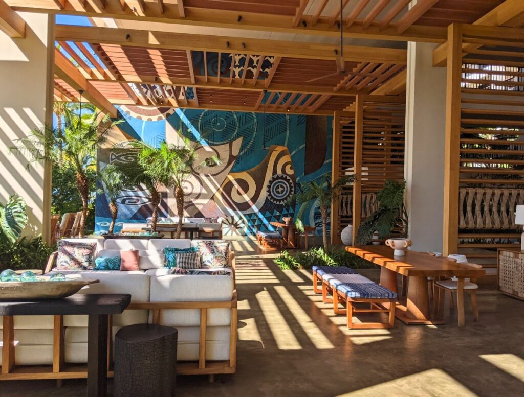Open air resort lounge with wooden beams, tropical plants, and colorful Hawaiian style mural artwork on the wall. Relaxing in stylish resort lounges and admiring local art is one of the laid back things to do Waikoloa between island adventures.
