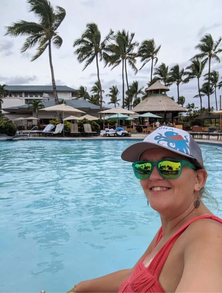 Woman smiling at the camera while relaxing in a large resort pool surrounded by tall palm trees and lounge chairs in Waikoloa. Enjoying resort pools and tropical scenery is one of the relaxing things to do Waikoloa on a Big Island vacation.
