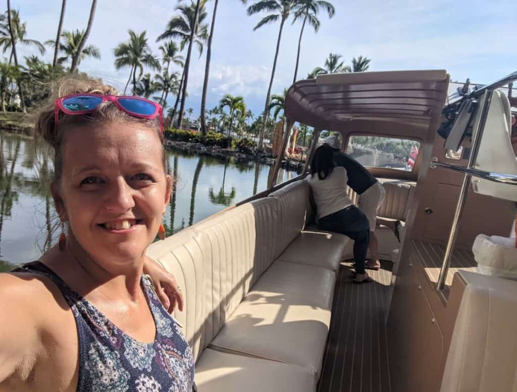 Woman taking a selfie while sitting in a small open air boat cruising through calm lagoons lined with palm trees at a Waikoloa resort. Taking a relaxing boat ride through the resort canals is one of the unique things to do Waikoloa visitors enjoy.