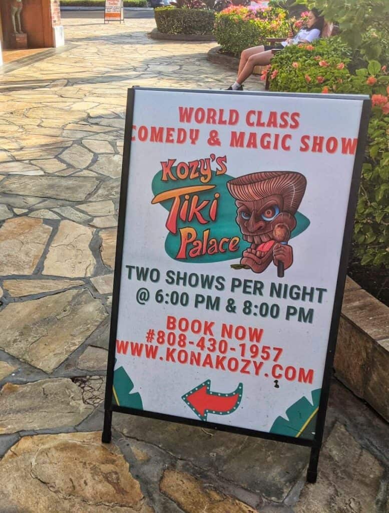 Sign on a stone walkway advertising "WORLD CLASS COMEDY & MAGIC SHOW Kozy’s Tiki Palace TWO SHOWS PER NIGHT @ 6:00 PM & 8:00 PM BOOK NOW #808-430-1957 WWW.KONAKOZY.COM
" with a tiki illustration. Attending this comedy and magic show is one of the unique evening things to do Waikoloa visitors can enjoy nearby.