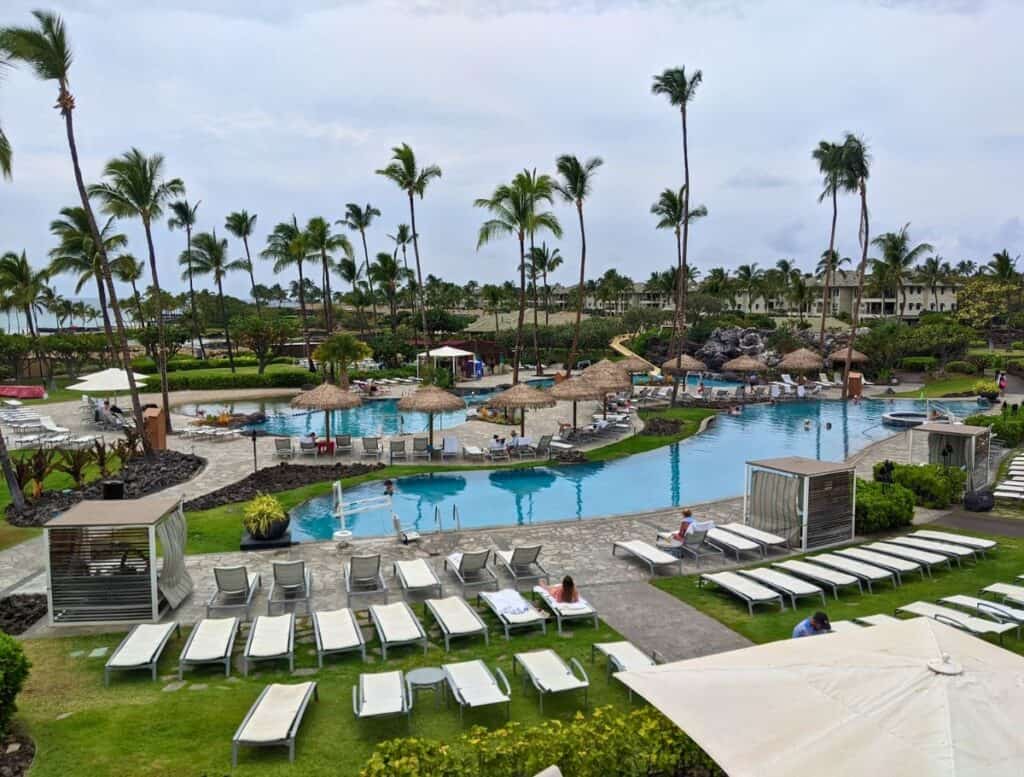 Large tropical resort pool complex with palm trees, lounge chairs, and thatched umbrellas overlooking the Waikoloa coastline. Relaxing at expansive resort pools like this is one of the classic things to do Waikoloa on a Big Island vacation.