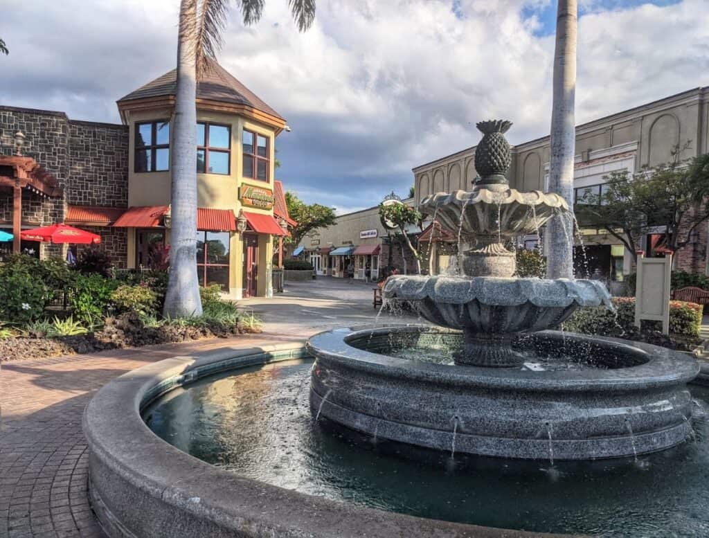 Stone fountain with a pineapple sculpture at the center surrounded by palm trees and shops at an open air shopping village in Waikoloa. Visiting local shopping areas and restaurants around the resort is one of the relaxing things to do Waikoloa visitors enjoy between beach and adventure activities.