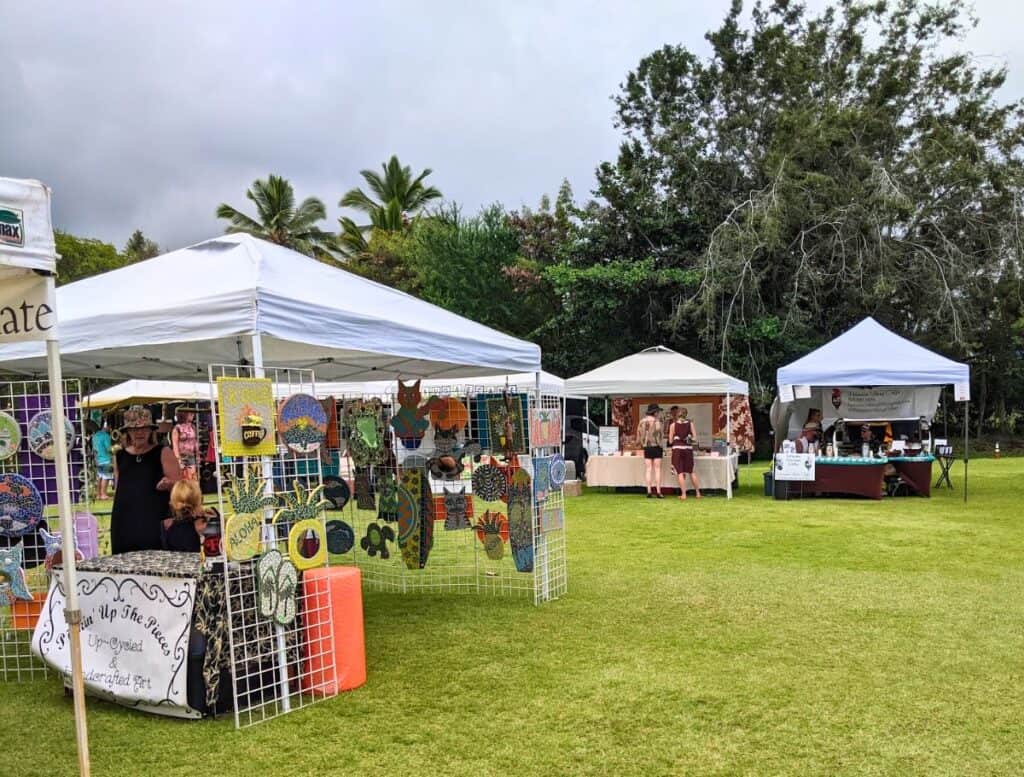 Outdoor farmers market on the Big Island with white canopy tents set up on a grassy field. Vendors display colorful handmade art and goods while shoppers browse, illustrating a typical stop when planning a big island farmers market schedule.