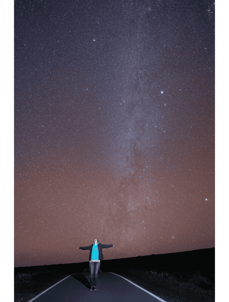 Person standing on a quiet road at night with arms outstretched beneath a sky full of stars and the Milky Way clearly visible above in Hilo Hawaii. Stargazing in dark sky areas offers a peaceful and awe inspiring experience and is one of the unique things to do in Hilo Hawaii.