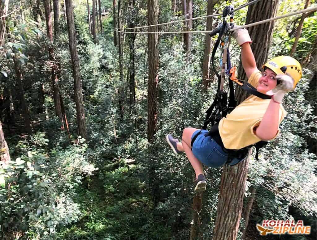 Person wearing a yellow helmet and harness rides a zipline high above a dense green forest while smiling and holding the cable. A logo in the corner reads "Kohala Zipline". Outdoor adventure activity through the treetops near Hawi.