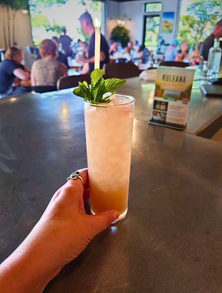 my hand holding a Kuleana Rum Shack mai tai with mint sprig in high ball glass and pink tinted drink, not dark like a normal mai tai.
