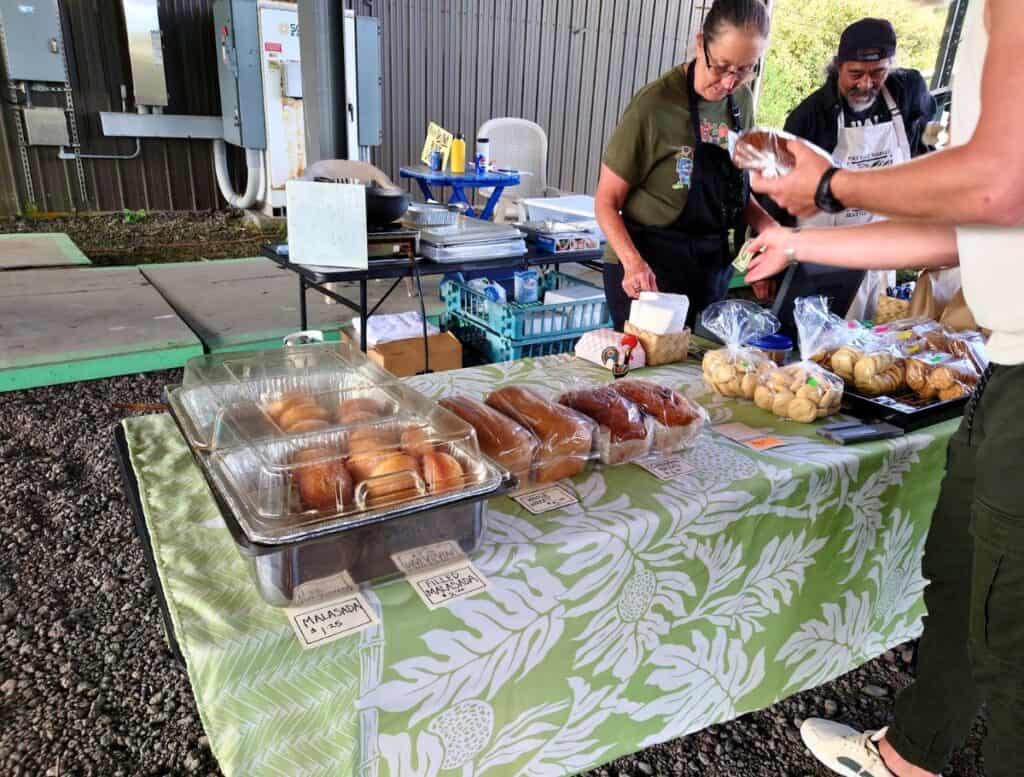Vendors sell fresh baked goods including bread rolls and packaged cookies at a local Big Island farmers market stall while a customer pays. Handwritten signs read "Malasada 1.25" and "Filled Malasada" showing typical food options to expect when following a big island farmers market schedule.