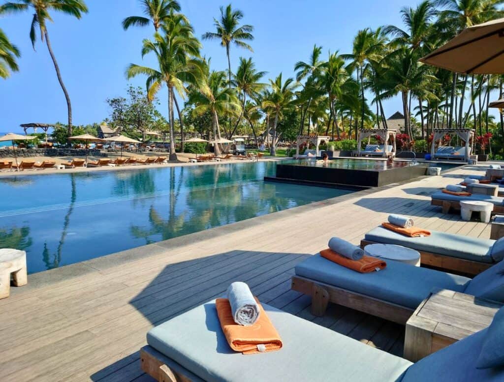 Tropical resort pool surrounded by tall palm trees with lounge chairs and rolled towels arranged along a wooden deck. Cabanas and umbrellas sit beside the water in a sunny oceanfront setting often highlighted in a Big Island travel guide.