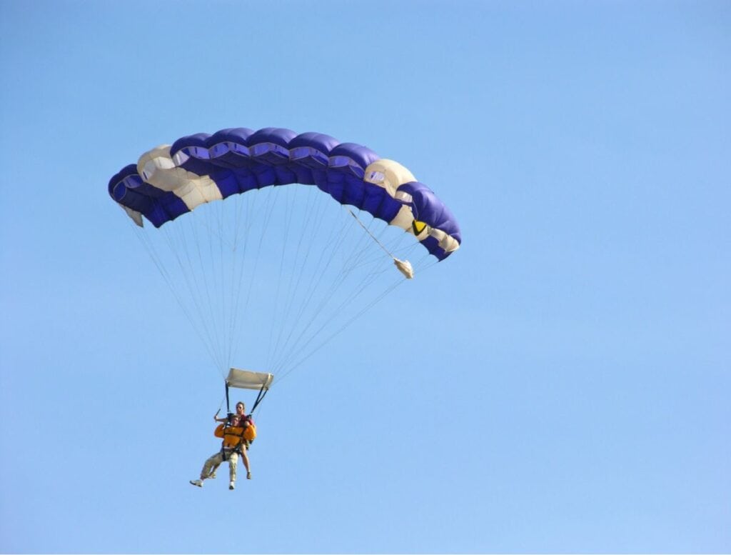 Two people tandem skydiving beneath a blue and white parachute high in a clear blue sky. Adventure activity on the Big Island that thrill seekers may add to their list of things to do Hawi while exploring the island.