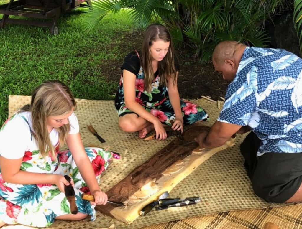 two girls sitting on woven mats taking Carving lessons at Aloha Adventure Farms led by a man teacher