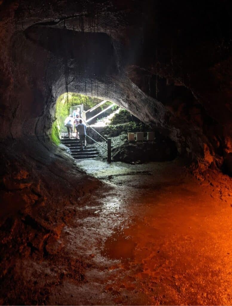 Visitors climbing steep stairs inside a dimly lit lava tube cave, with bright artificial lights highlighting the rock walls