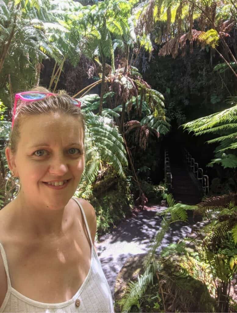 me wearing pink sunglasses on my head, with lush ferns and a dark thurston lava tube entrance in the background