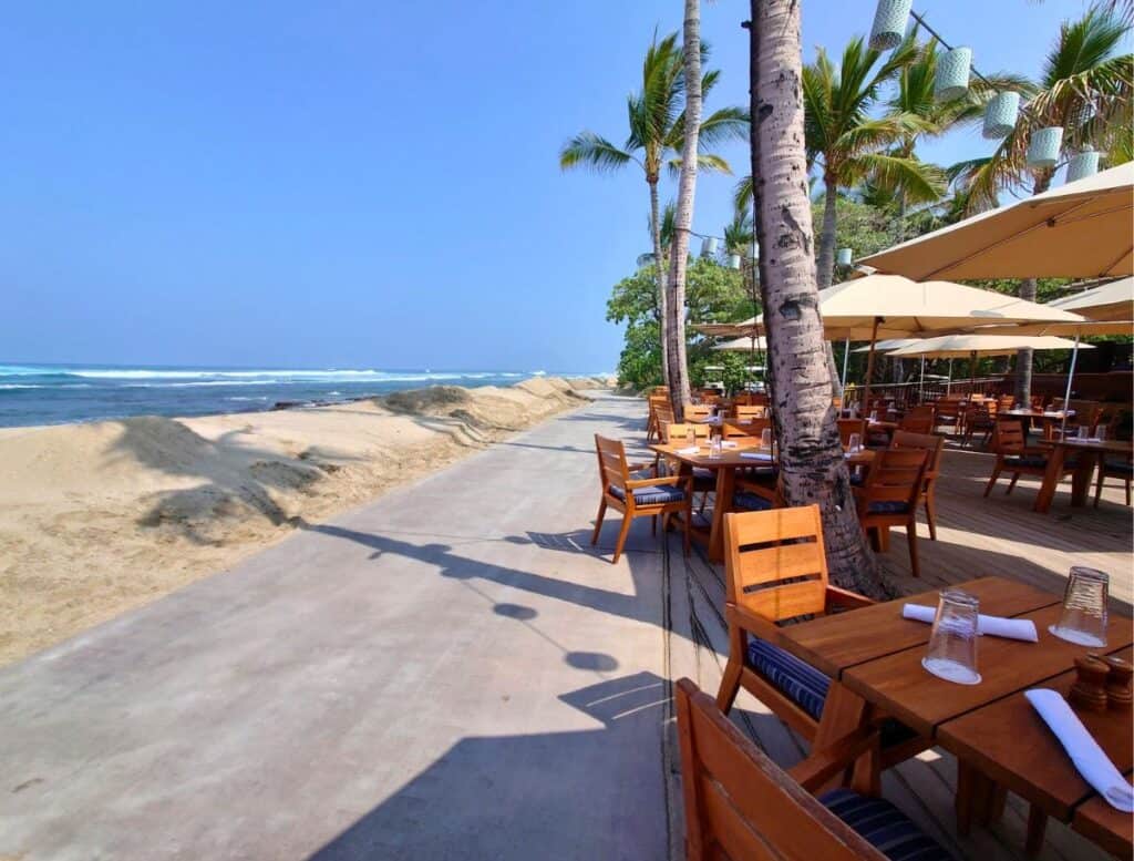 outdoor restaurant with wooden tables under umbrellas and palm trees. paved walking path runs next to it with white sand and ocean on the other side