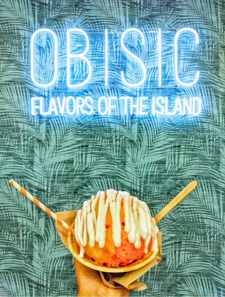 obisic bowl of fruity shave ice and drizzled haupia cream on top under a neon sign that reads OBISIC Flavors of the Island