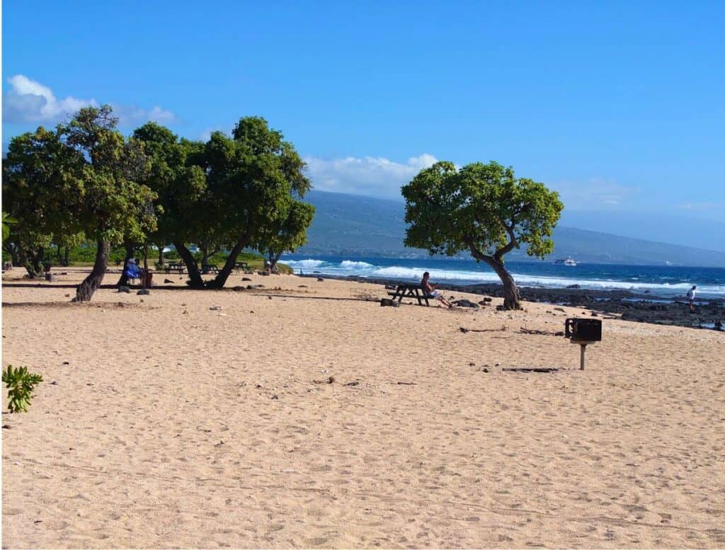 Old Kona Airport Beach with white sand, lava pads, people sitting at picnic tables under trees and lone bbq