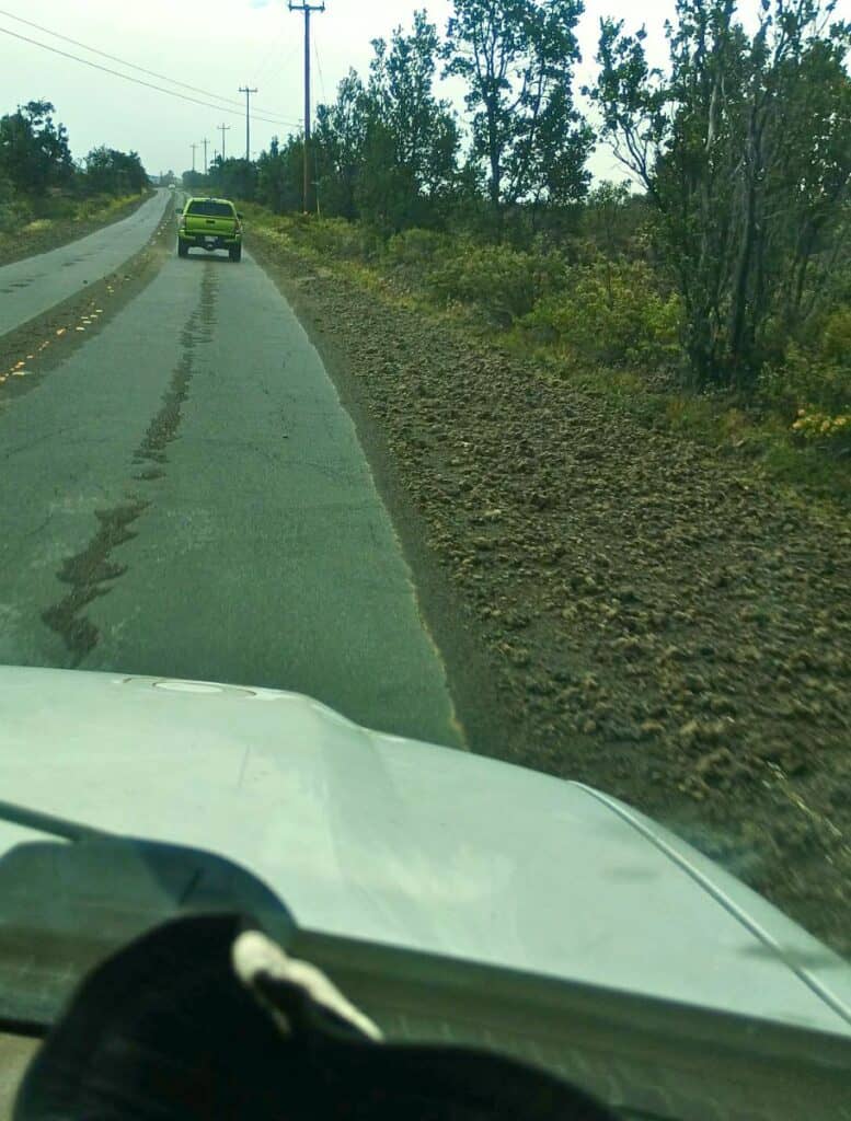 View from inside a vehicle driving along a rural Big Island road where the shoulder is covered with loose volcanic rocks and tephra. A green truck drives ahead while scattered lava fragments along the roadside show conditions that can contribute to tephra damage car rental risks during Big Island travel.