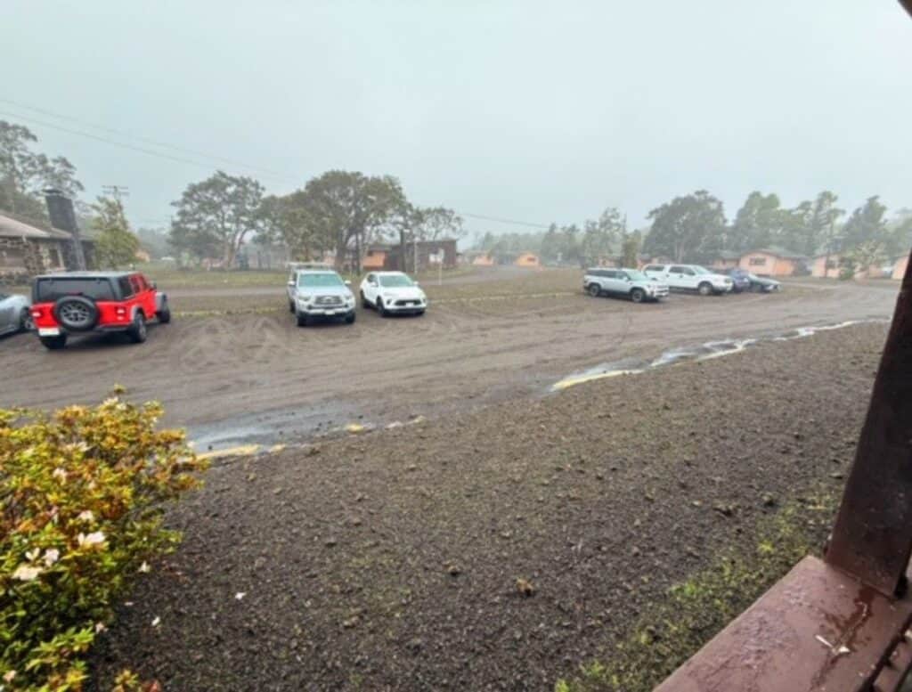 A muddy parking area coated in wet volcanic ash surrounds several parked cars under a gray hazy sky near small buildings and trees. The damp conditions show how tephra damage rental car exteriors by sticking to surfaces and creating abrasive grime when driven through.