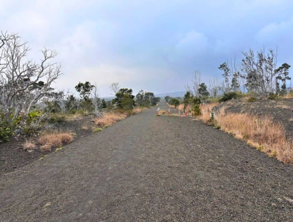 A long unpaved road covered in dark volcanic gravel stretches through dry grass and scattered trees under a cloudy sky. The loose tephra surface shows how tephra damage rental car tires and paint when driving along rough volcanic roads.